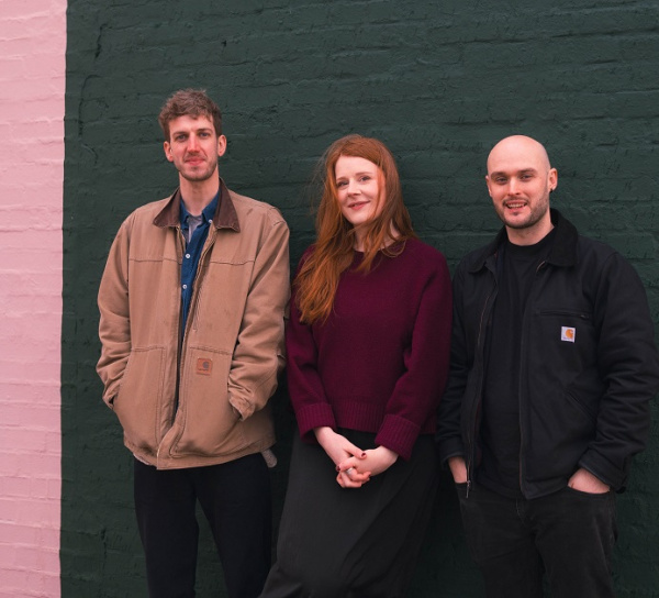 Zwei Musiker und eine Sängerin stehend vor einer Wand.