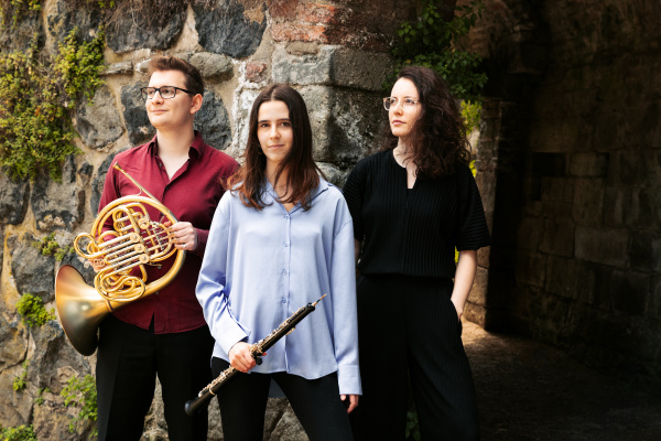Die drei jungen Musiker:innen stehen vor einem Gebäude aus, mit Mauerfugenvegetation bewachsenen Natursteinen.